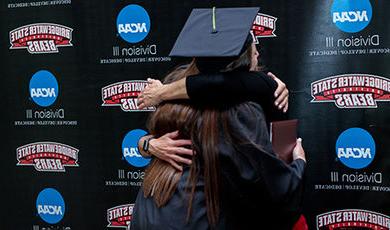 A graduate hugs a coach in front of a BSU athletics backdrop.