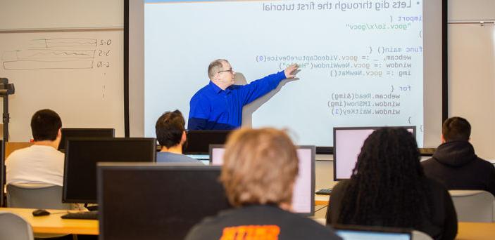 A faculty member teaches pointing to a projector screen as students with open laptops look on  